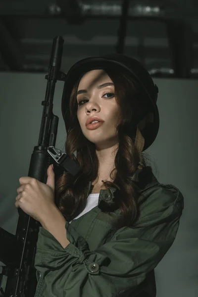 Beautiful girl posing in military helmet with rifle, on grey with shadows — Stock Photo