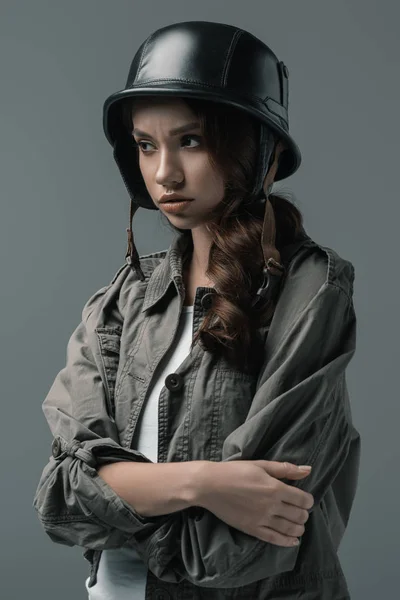 Belle fille inquiète posant dans un casque militaire avec les bras croisés, isolé sur gris — Photo de stock