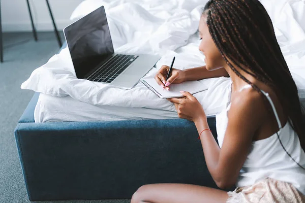 Жінка з ноутбуком в ліжку пише в блокноті — стокове фото