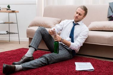 Handsome man sitting on a floor in the living room with smartphone and bottle of beer clipart