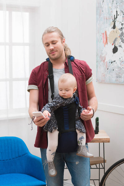 Father standing with son in baby sling and looking at smartphone