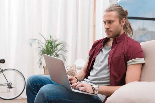自宅のソファの上に座って ノート パソコンを見ているハンサムな男  — 無料ストックフォト