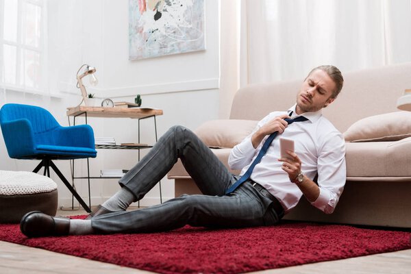 Бизнесмен сидит на ковре, развязывает галстук и смотрит на смартфон
