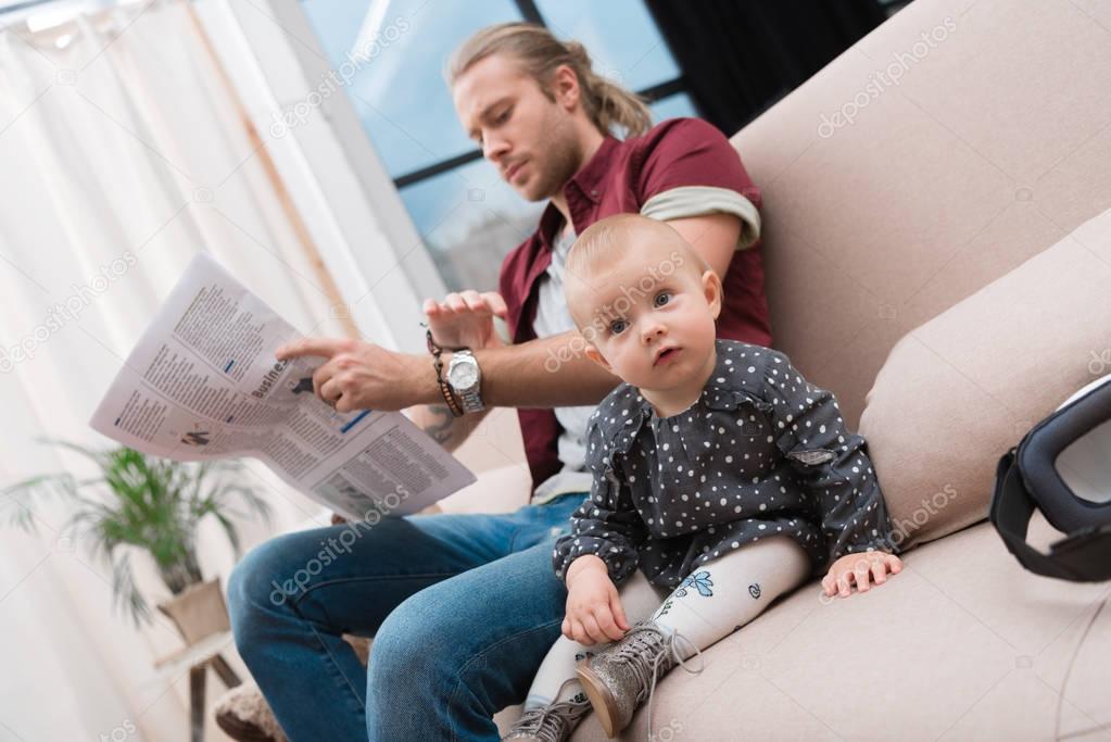 little baby girl sitting on sofa while her father reading newspaper at home