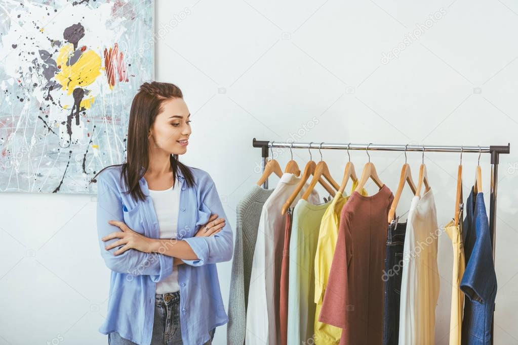 smiling designer with crossed hands looking at clothes  