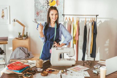smiling seamstress posing with scissors at working place and looking at camera clipart