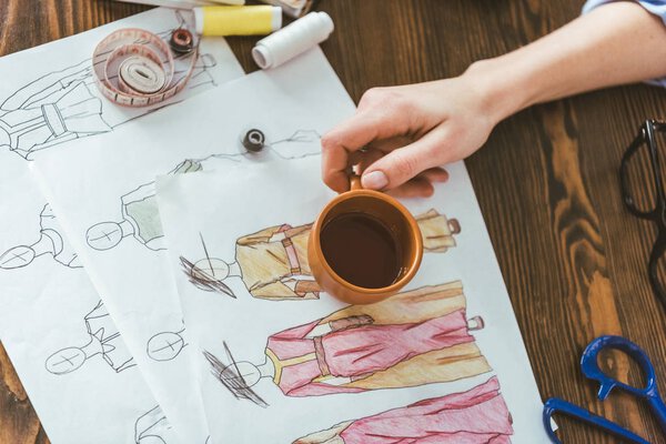 cropped image of seamstress holding cup of tea above sketches