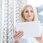 Professional female ophthalmologist working with digital tablet in optics with glasses on shelves