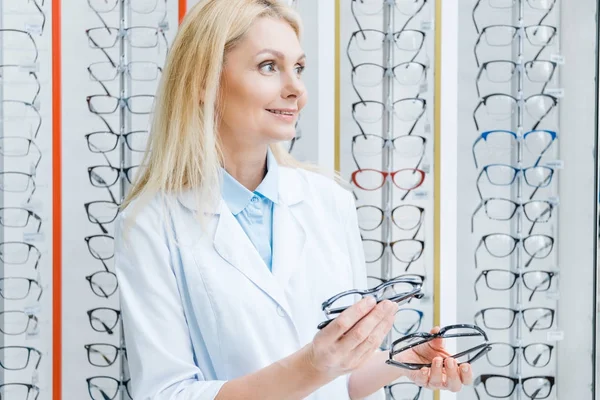 Χαμογελαστά Γυναικεία Οφθαλμίατρο Κρατώντας Γυαλιά Και Στέκεται Στην Οπτική — Δωρεάν Φωτογραφία