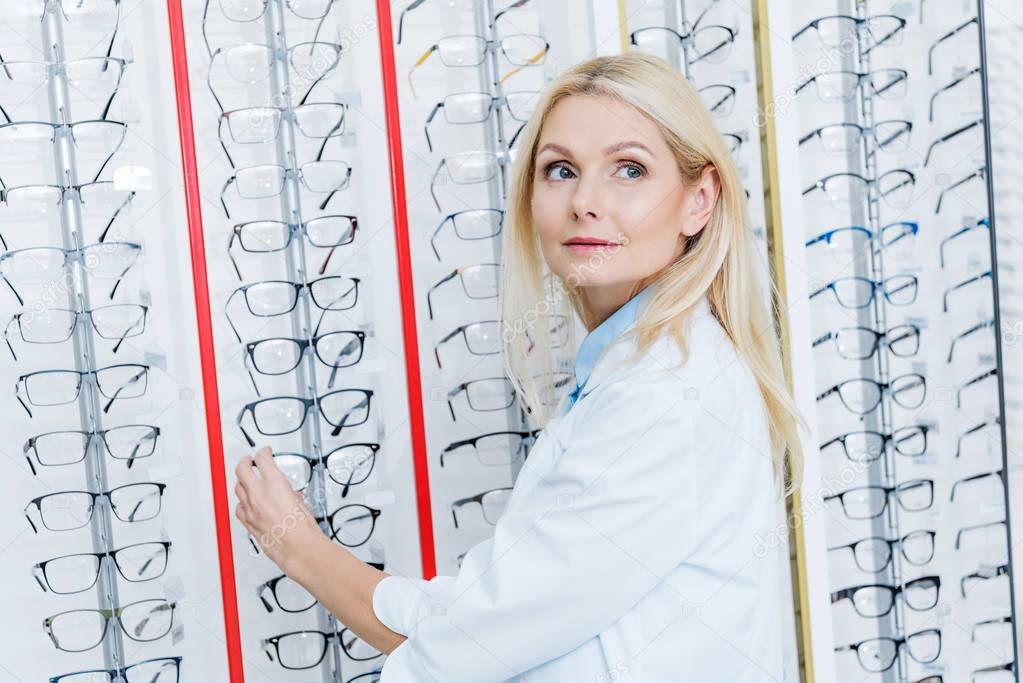 beautiful female ophthalmologist standing in optics with glasses on shelves