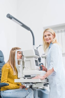 professional female optometrist examining patient through slit lamp in clinic clipart