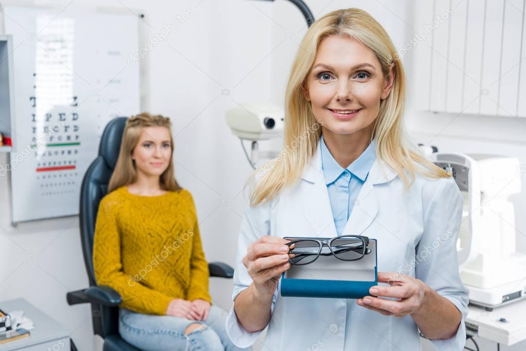 optician in white coat holding eyeglasses while patient sitting behind