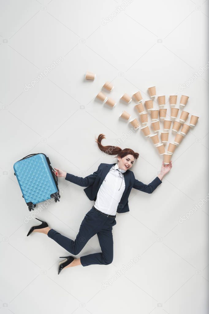 overhead view of cheerful businesswoman with suitcase and disposable cups isolated on grey