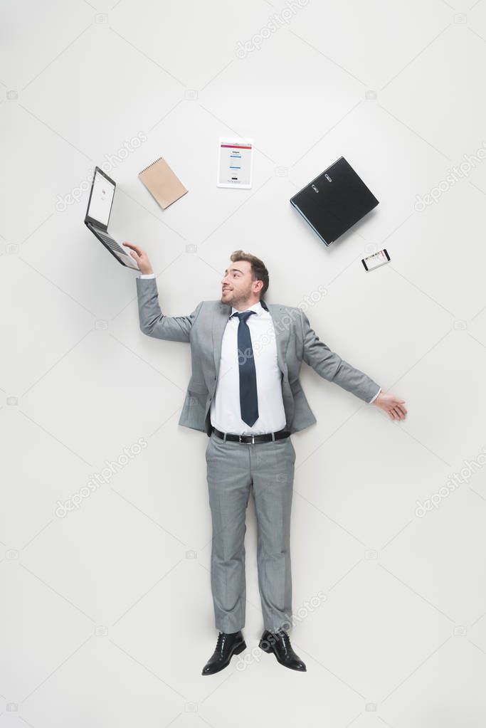 Overhead view of businessman with office supplies above head using laptop isolated on grey