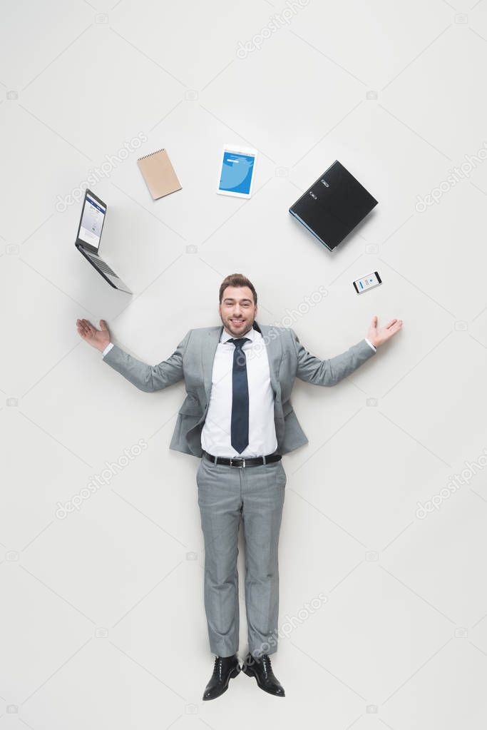 Overhead view of smiling businessman with outstretched arms and office supplies above head looking at camera isolated on grey