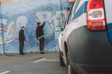 back view of vandals painting graffiti on wall, police car on foreground clipart