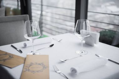 close up view of arranged cutlery, empty wineglasses and menu on table with white tablecloth in restaurant clipart