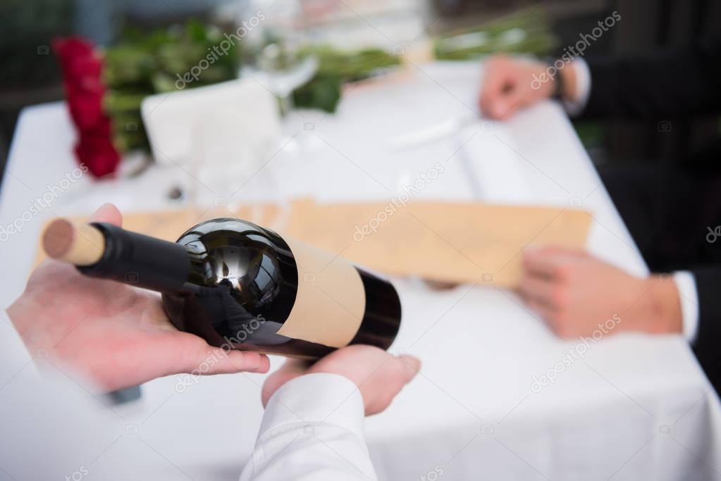 selective focus of waiter showing bottle of wine to visitor at table in restaurant