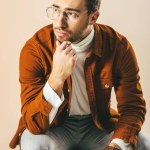 Retrato de hombre elegante reflexivo sentado en una silla de madera y mirando hacia otro lado aislado en beige