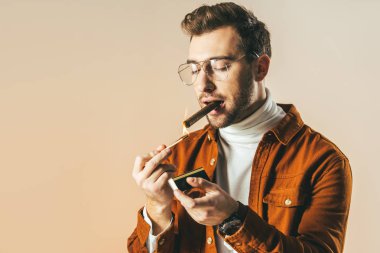 portrait of stylish man in eyeglasses lighting up cigar isolated on beige