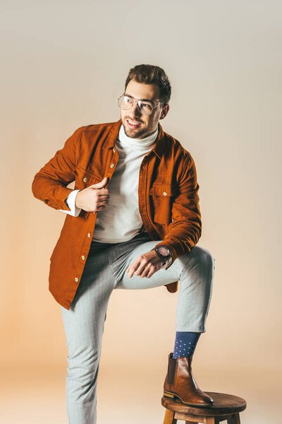 cheerful handsome man with one leg on chair looking away isolated on beige