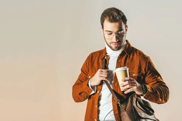 コーヒーに手で孤立したベージュの使い捨てカップを見てバッグを持つファッショナブルな男の肖像 — ストック写真