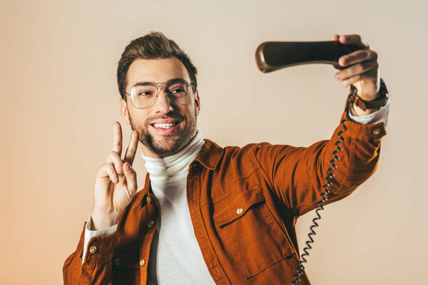 portrait of smiling man showing peace sign while pretending taking selfie in telephone tube isolated on beige