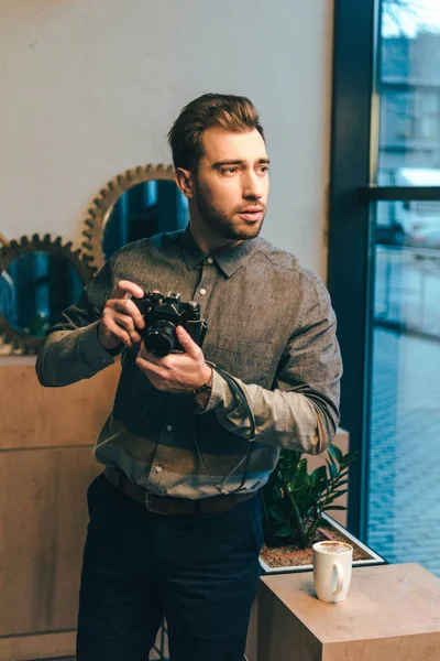 portrait of thoughtful man with photo camera standing at window in cafe