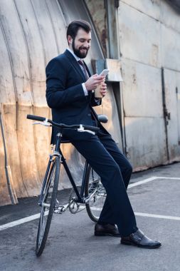 smiling businessman with coffee to go using smartphone while leaning on bicycle on street clipart