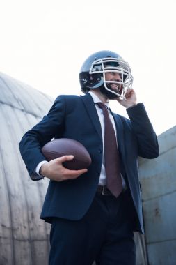 low angle view of young businessman in suit and rugby helmet holding ball in hands on street clipart