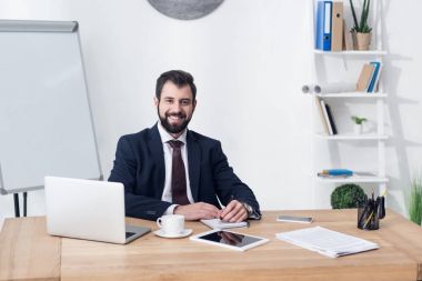 İşyerinde ofiste otururken kameraya bakarak gülümseyen işadamı portresi