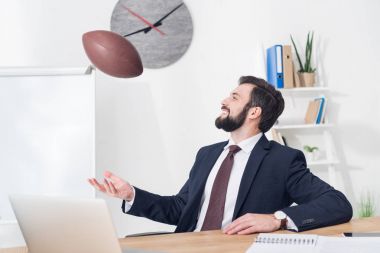 portrait of businessman throwing rugby ball at workplace with laptop in office clipart