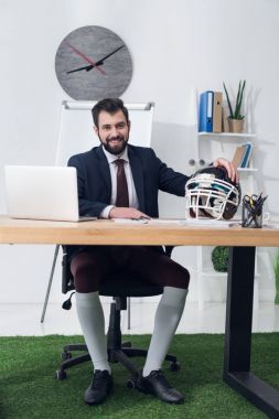 young businessman sitting at workplace with rugby helmet in office clipart