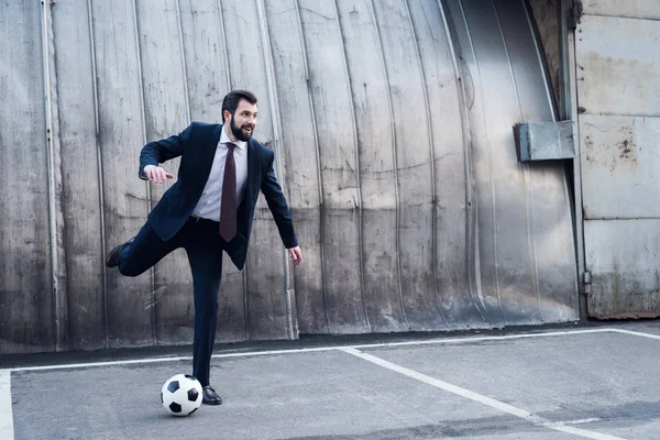 young smiling businessman in suit playing soccer on street
