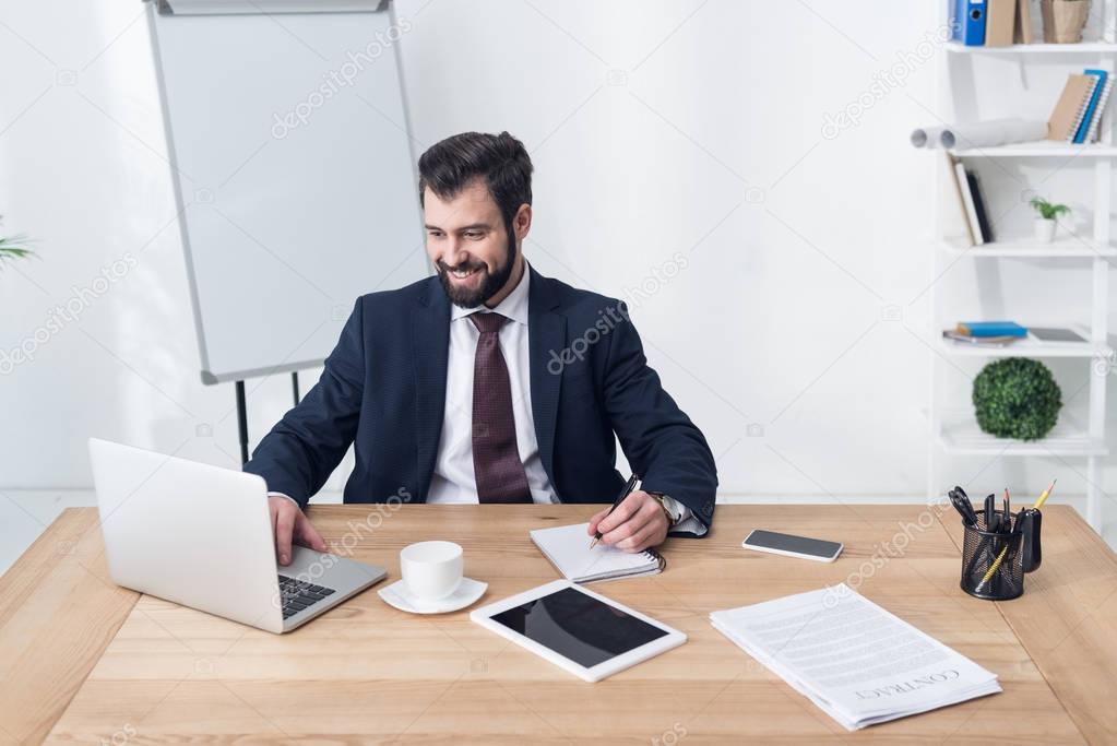 portrait of smiling businessman typing on laptop and making notes in notebook at workplace in office