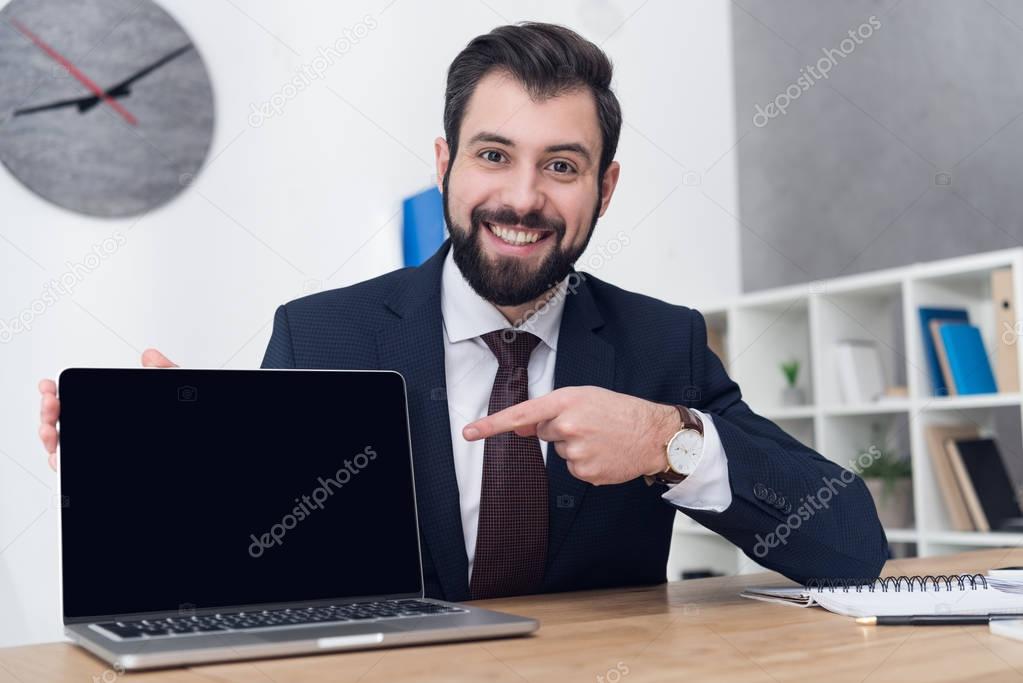 portrait of cheerful businessman pointing at laptop at workplace in office