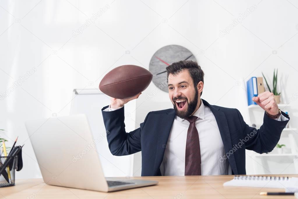 portrait of excited businessman with rugby ball at workplace with laptop in office
