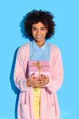 portrait of cheerful african american woman with gift in hands against blue wall