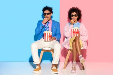 young african american couple sitting on chairs and eating popcorn against pink and blue wall