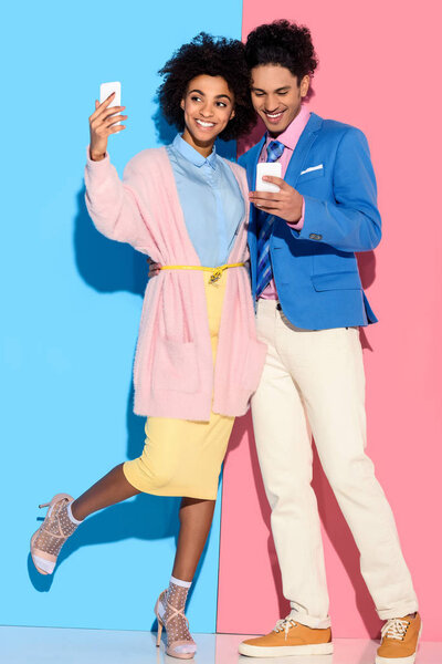Young african amercian girl taking selfie and hugging stylish guy who looks at his phone on pink and blue background 