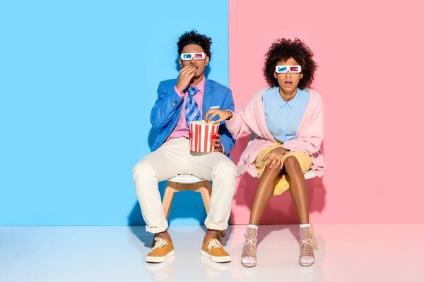 young african american couple sitting on chairs and eating popcorn against pink and blue wall