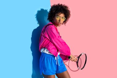 Genç parlak Afro-Amerikan kız tenis raketi pembe ve mavi arka plan üzerinde sallanır. 