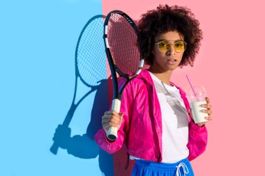 Genç parlak Afro-Amerikan kız tenis raketi ve plastik bardak içki ile pembe ve mavi arka plan üzerinde tutan 