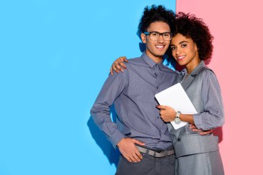 Young african amercian businesswoman holding tablet and hugging stylish businessman on pink and blue background  clipart