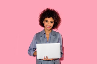 Young african amercian smiling businesswoman holding laptop on pink background  clipart