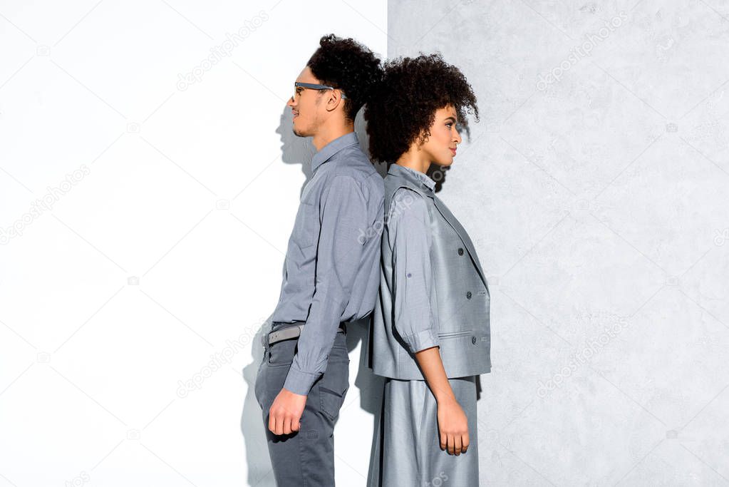 Young african amercian smiling couple in grey suits standing back to back on grey and white background 