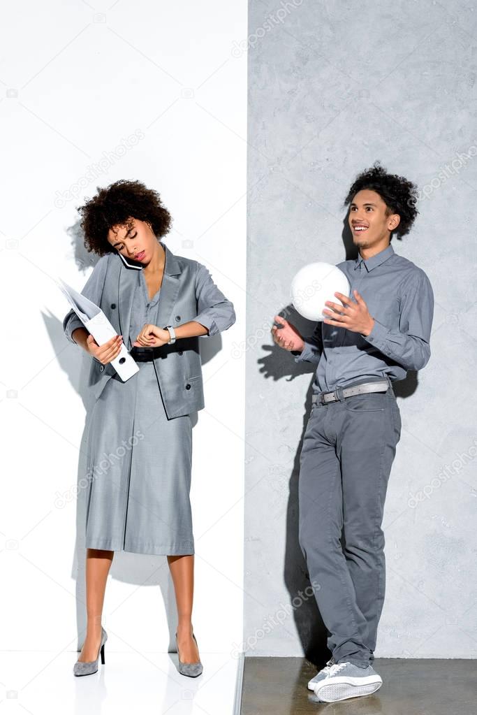 Young african amercian busy businesswoman holding folder and talking on phone while relaxed guy playing with ball on grey and white background 