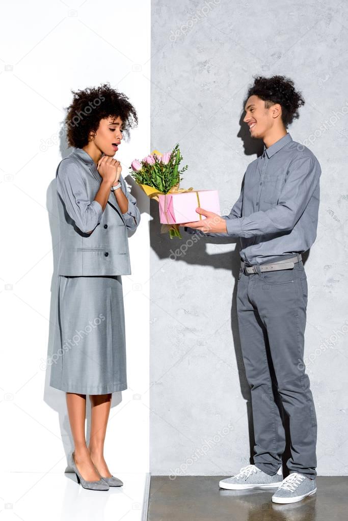 Young african amercian man gives girl a bouquet of flowers and gift on grey and white background 