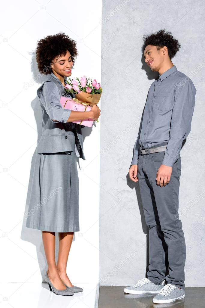 Young african amercian girl holding bouquet of flowers and gift presented by smiling man on grey and white background 