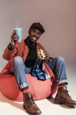 Hamburger yeme ve Studio fasulye çanta sandalyede otururken kağıt bardak içme mutlu genç Afro-Amerikan adam